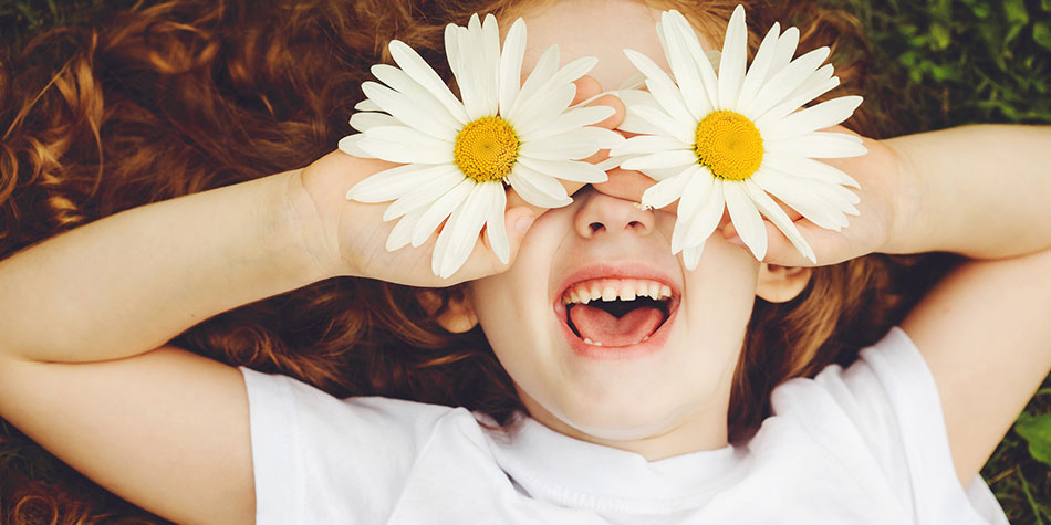 Little girl smiling, laying on grass holding spring daisies over her eyes
