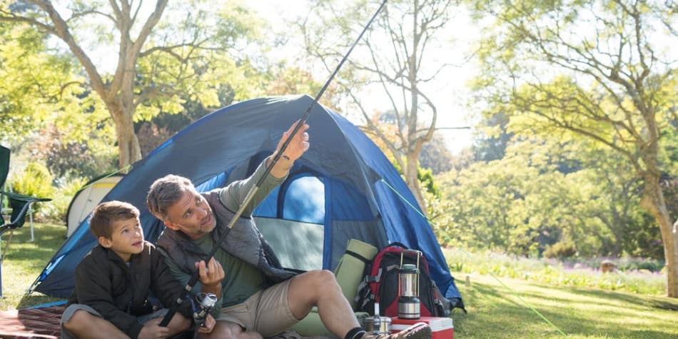 A father and son adjust a fishing pole outside of a tent while camping
    outside.