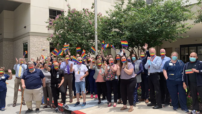 hospital staff and community members stand together outside on sidewalk wearing masks holding raised pride flags in front of flag pole with pride flag outside of hospital