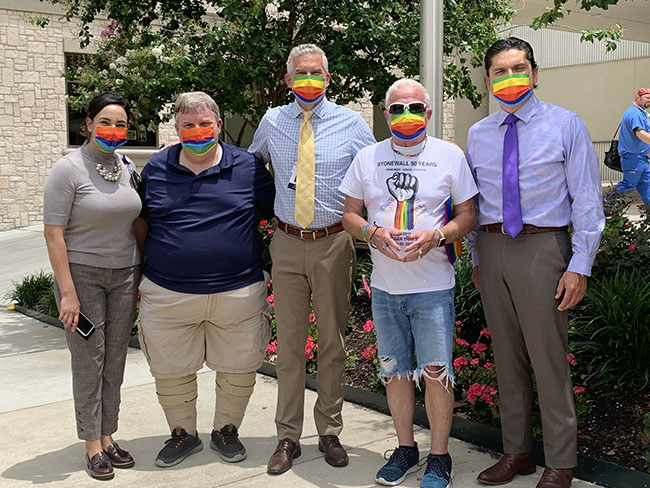 small group of hospital staff stand with member of pride center san Antonio wearing masks in front of flag pole outside hospital