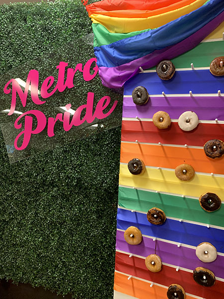 rainbow wall installation with pegs holding donuts with rainbow flag draped over top leaning against greenery wall