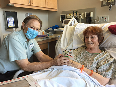 Older woman lying in hospital bed smiling holding man’s hand wearing face mask