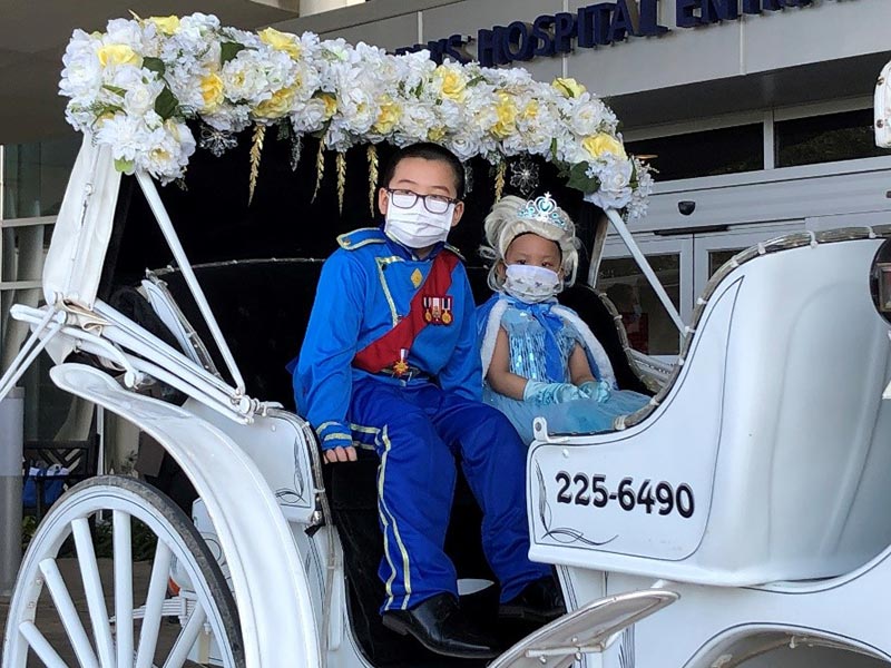 little girl and boy dressed up as Cinderella and Prince Charming sitting in white carriage decorated with flowers outside hospital entrance